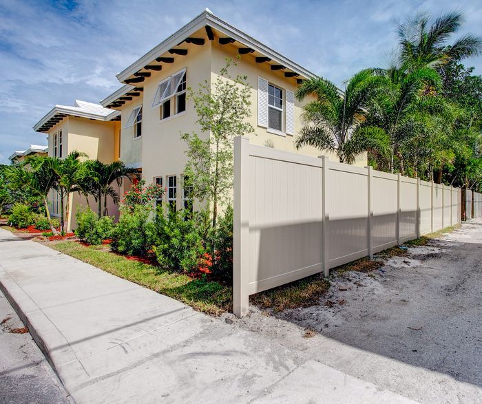 Charming two-story yellow building surrounded by lush greenery and palm trees, featuring a modern white fence for privacy in a sunny neighborhood.