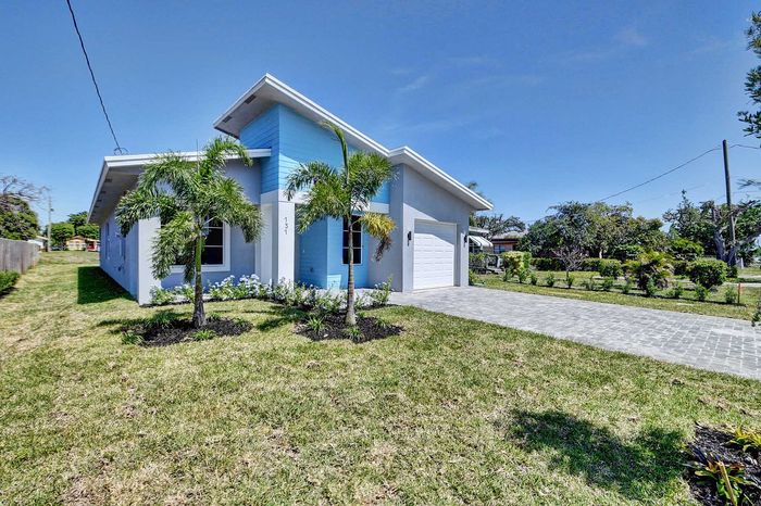 Charming modern home with sleek blue accents and palm trees, nestled in a lush green landscape, featuring a spacious driveway and manicured lawn.