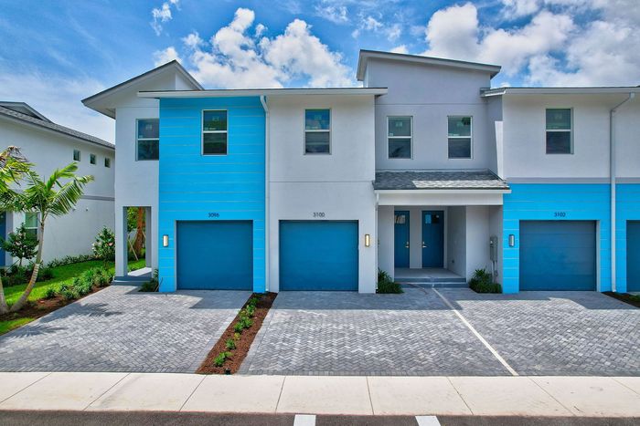 Contemporary townhouse with vibrant blue accents, featuring individual garages and lush greenery under a clear sky.