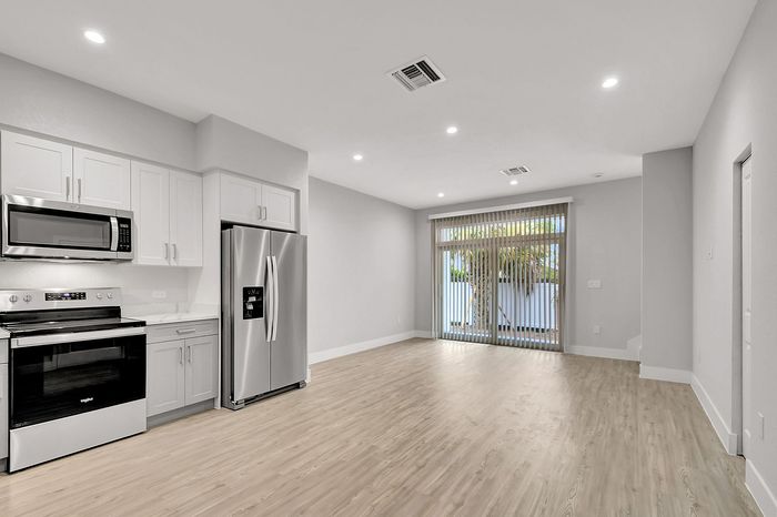 Modern open-concept living and kitchen area with sleek stainless steel appliances, wooden flooring, and large sunlit windows.