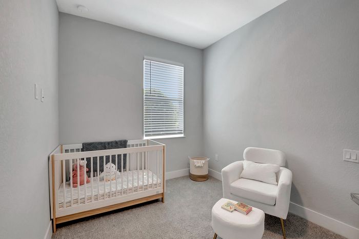 Charming nursery with cozy crib, plush seating area, and soft carpet accented by natural light for a serene ambiance.