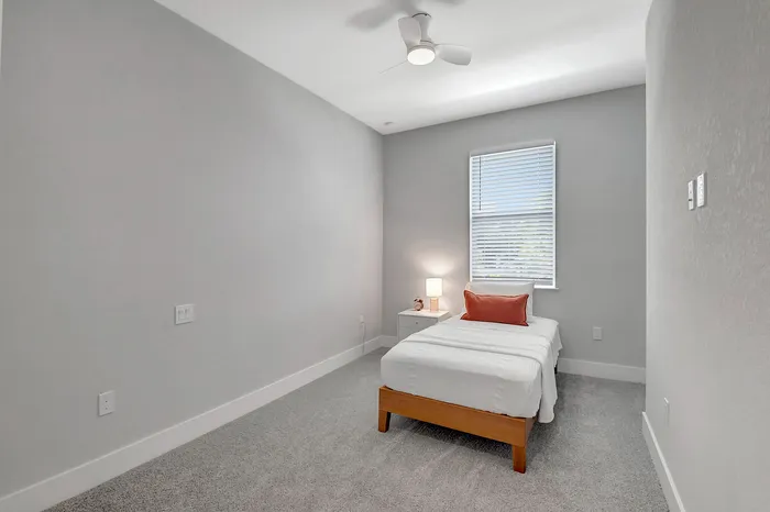 Bright and cozy bedroom with a modern ceiling fan, featuring a single bed dressed in crisp white linens and a plush orange accent pillow, inviting relaxation.