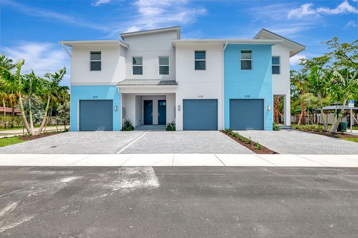 Modern duplex with tropical landscaping featuring vibrant blue accents and spacious driveways under a sunny sky.