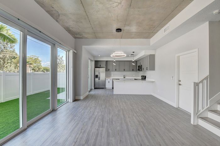 Modern open-plan kitchen and living area with sleek flooring, stylish pendant lighting, and expansive glass doors leading to a private outdoor space with lush greenery.