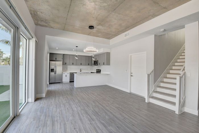 Modern open-concept living area with sleek kitchen, featuring stainless steel appliances, stylish pendant lighting, and textured ceiling, adjacent to a bright space with large sliding glass doors and a contemporary staircase.