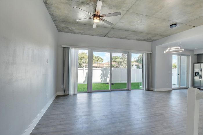 Modern living space with sleek wooden flooring, expansive glass doors, and stylish ceiling fan, leading to lush green outdoor area.
