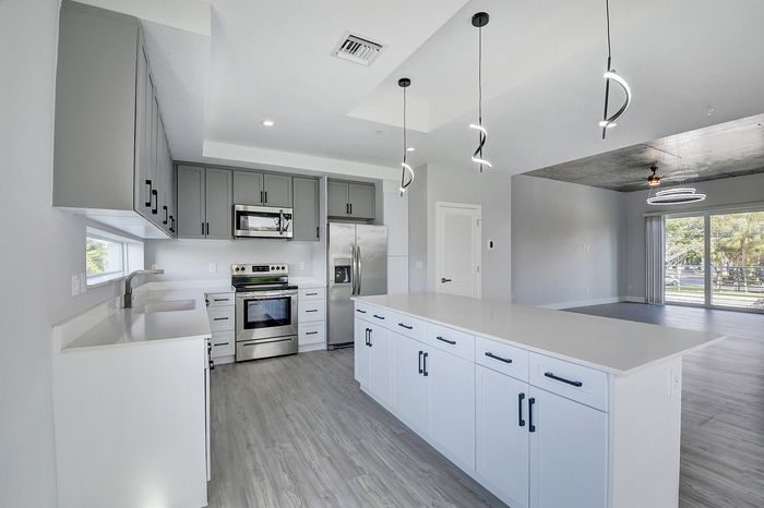Modern kitchen with sleek grey and white cabinetry, stainless steel appliances, and elegant pendant lighting opening to a spacious, light-filled living area.