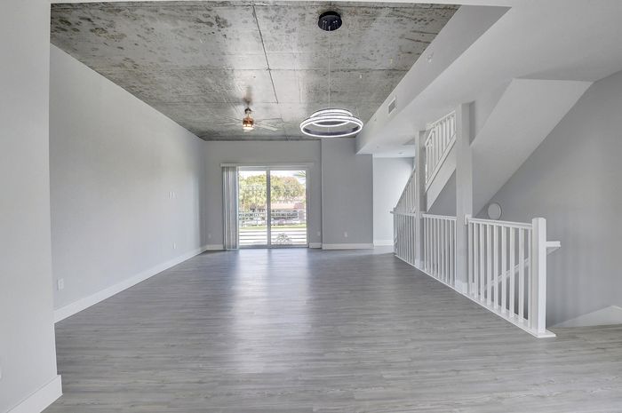 Contemporary open-plan living space with sleek gray flooring and industrial-style concrete ceiling, complemented by modern lighting and abundant natural light from large glass doors.
