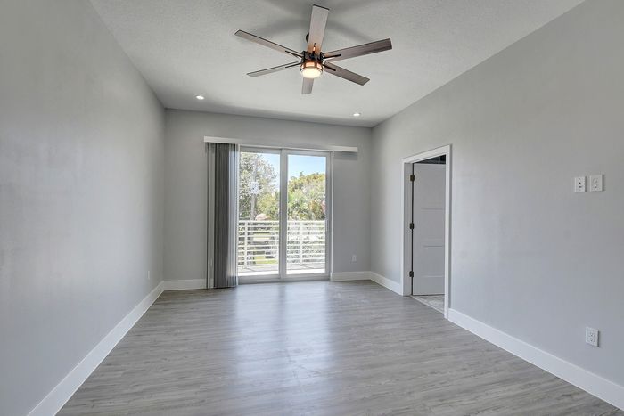 Bright and spacious room featuring elegant wood flooring, a modern ceiling fan, and sliding glass doors leading to a sunlit balcony with a serene view.