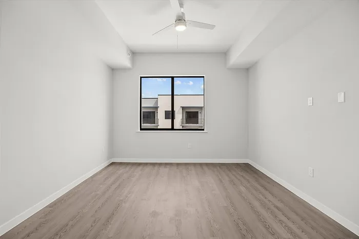 Bright, airy room with sleek wooden flooring, modern ceiling fan, and large window offering urban views.