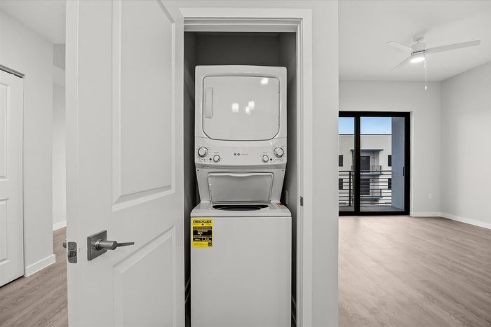 Contemporary apartment view featuring an in-unit washer and dryer set in a sleek closet, adjacent to a bright living space with stylish flooring and a balcony access.