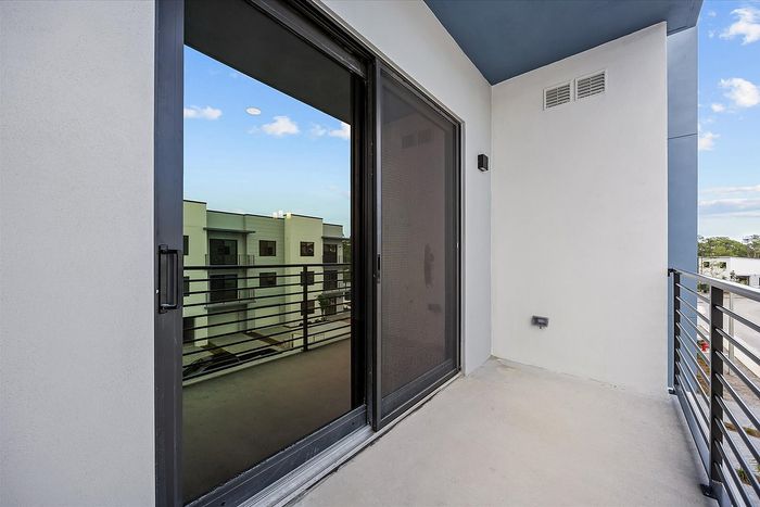 Modern apartment balcony with sleek glass sliding door and stylish metal railing, offering clear views of neighboring buildings and a bright blue sky.