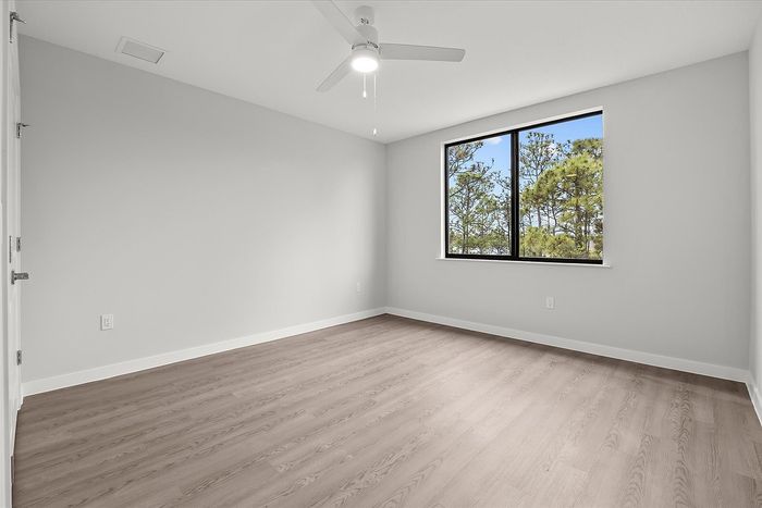 Bright and inviting bedroom with sleek light-colored flooring, modern ceiling fan, and a large window revealing a scenic tree view.