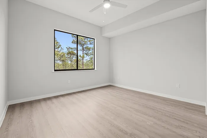 Bright, airy bedroom with large window overlooking scenic greenery, featuring elegant hardwood floors and modern ceiling fan.