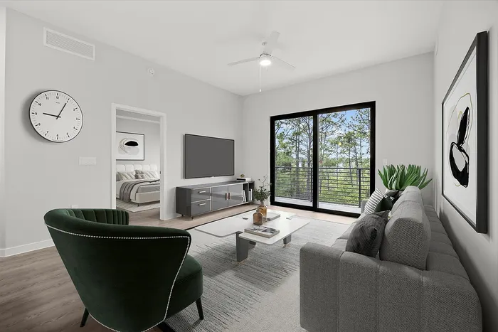 Modern living space with a cozy gray sofa, chic green armchair, and a sleek entertainment center. Large windows offer a serene view of lush greenery, and a stylish bedroom is visible through an open doorway.