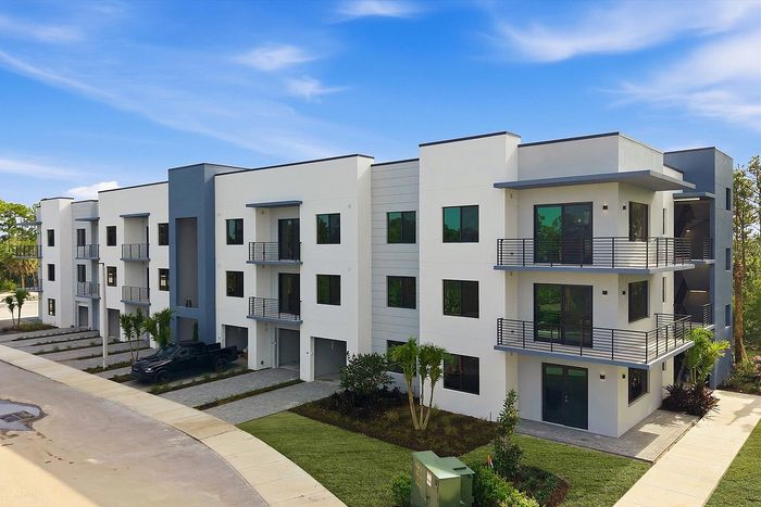 Modern apartment building with clean white facade, spacious balconies, and lush surrounding greenery under a vibrant blue sky.