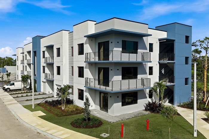 Modern three-story apartment building with stylish balconies, surrounded by lush landscaping under a clear blue sky.