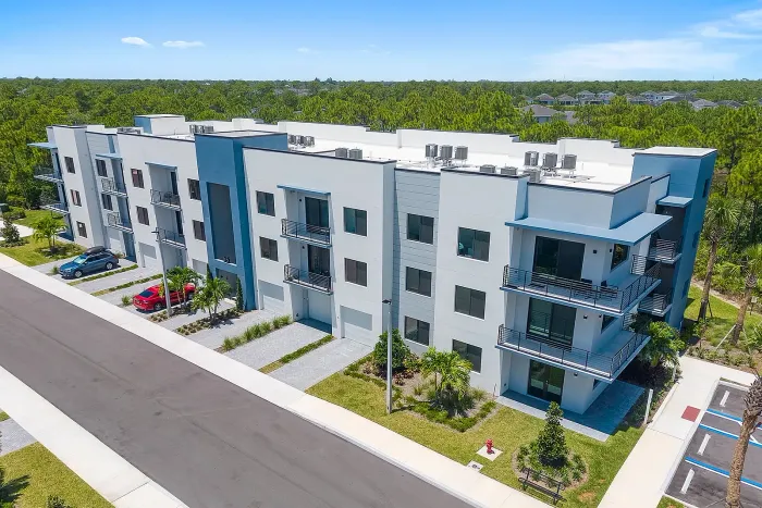 Aerial view of a modern three-story white and blue apartment building featuring private balconies, and paved driveways backed by a dense green forest