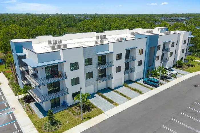 High-angle shot showing the side profile, corner balconies, and ground-level garages of a modern three-story residential complex under a clear blue sky.