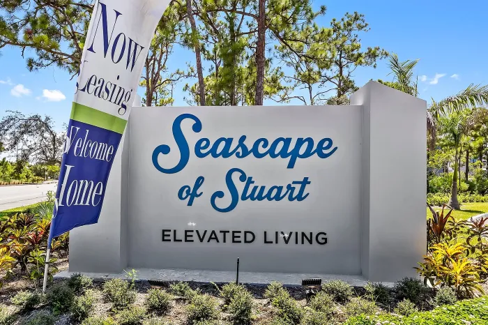 Ground-level entrance sign reading "Seascape of Stuart ELEVATED LIVING" in blue lettering on a grey monument wall, framed by tropical foliage and a "Now Leasing" feather banner.