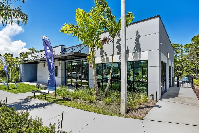 terior of a modern single-story community clubhouse featuring large floor-to-ceiling glass windows, grey paneled siding, palm trees, and a blue