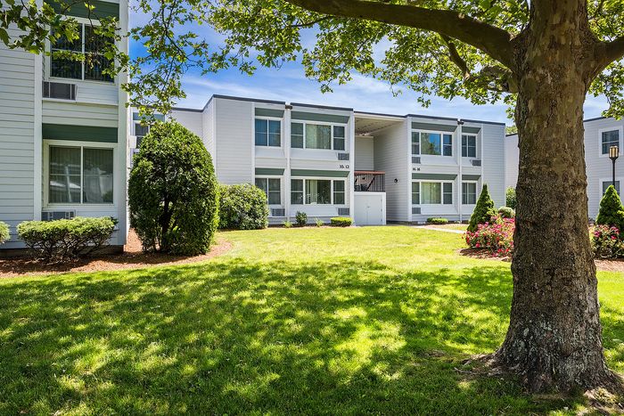 Charming apartment complex with bright white exterior, lush green lawn, and beautiful landscaping under a sunny blue sky.