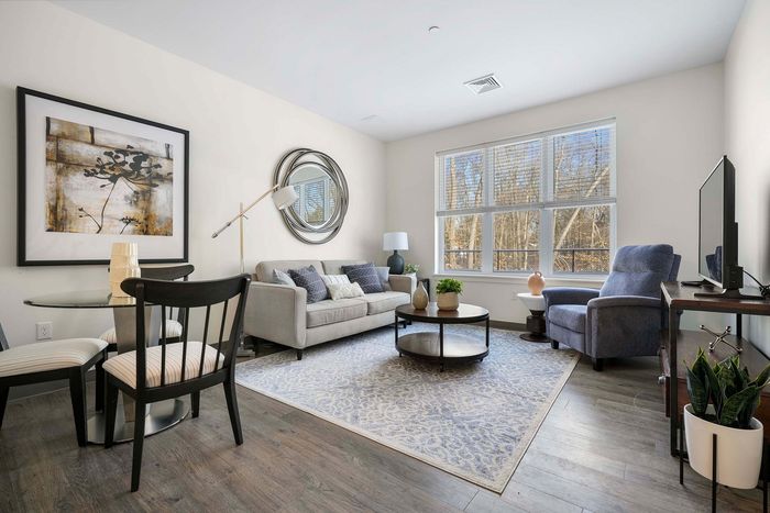 Bright and modern living room with elegant decor, featuring a cozy gray sofa, sleek round mirror, and stylish artwork. Large windows offer plenty of natural light, illuminating the chic furnishings and inviting atmosphere.