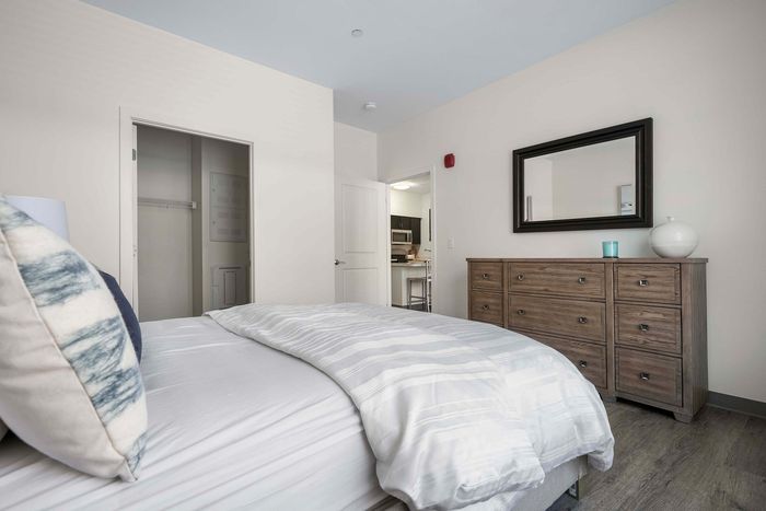 Inviting bedroom with sleek hardwood floors, modern dresser, plush bed, and a glimpse of a cozy kitchen.