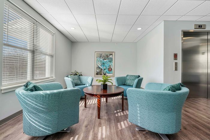 Inviting lounge area with elegant teal armchairs, a round wooden table, and abundant natural light from large windows, perfect for relaxation and conversation.