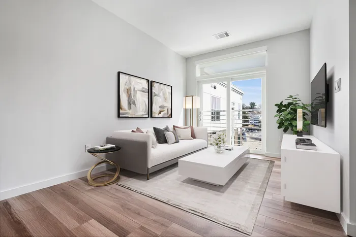Bright, modern living room featuring elegant wooden flooring, a plush sofa with decorative cushions, a sleek white coffee table, and natural light streaming through large glass doors.