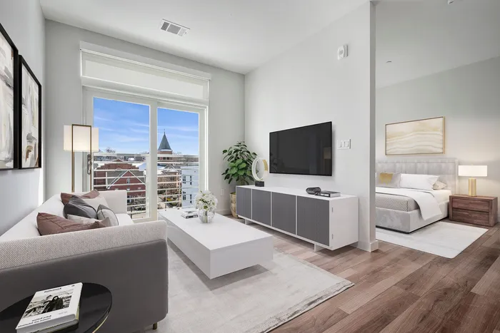 Modern apartment living room with stylish decor, large TV, and bright natural light from balcony. Cozy bedroom glimpsed through open doorway.