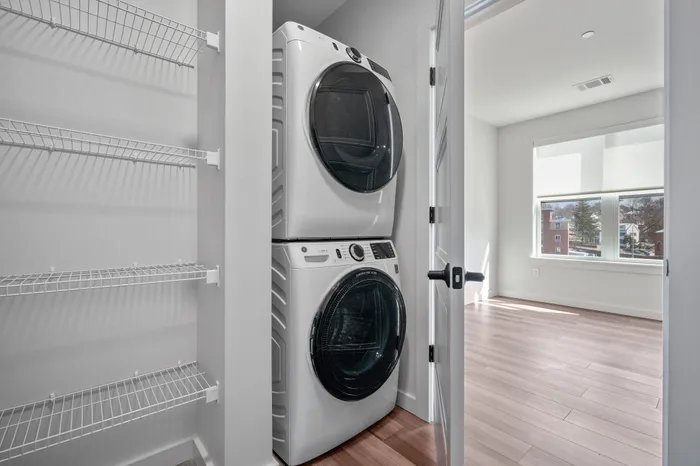 Bright laundry room with sleek stacked washer and dryer next to spacious shelving in a modern apartment.