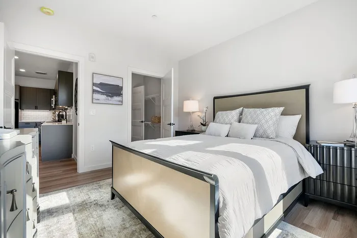 Bright and inviting bedroom with elegant decor, featuring a plush bed, modern furniture, and a view into a sleek kitchen with stylish cabinetry.