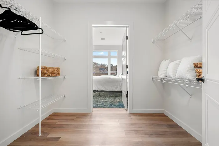 Bright walk-in closet leading to a cozy bedroom with large windows, featuring modern shelving and elegant wooden flooring.