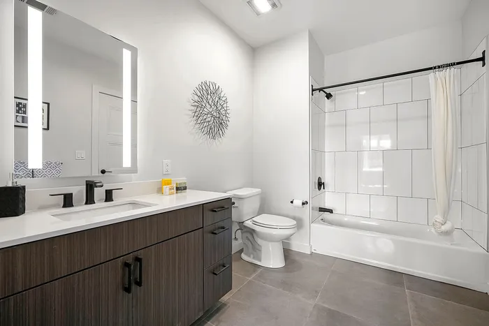 Modern bathroom with sleek dark wood vanity, large mirror with LED lighting, and chic wall art, featuring a spacious bathtub-shower combo with elegant tile work.
