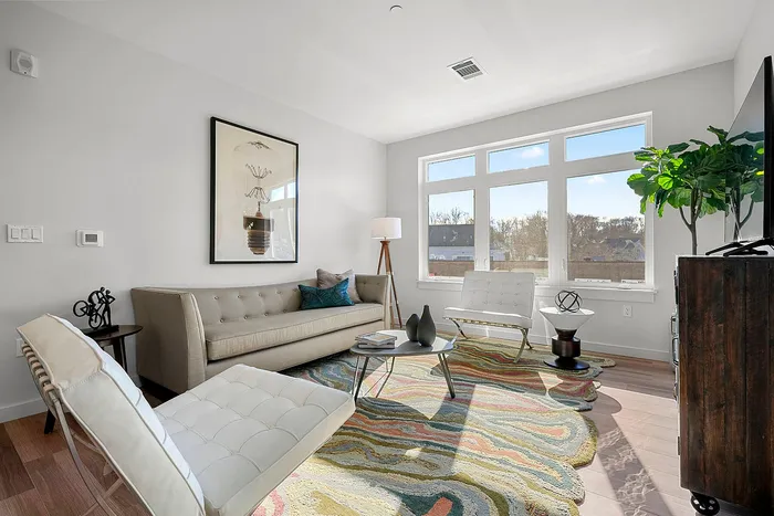 Bright, modern living room featuring chic decor, a plush beige couch, stylish armchairs, and a vibrant patterned rug, illuminated by natural light through large windows.