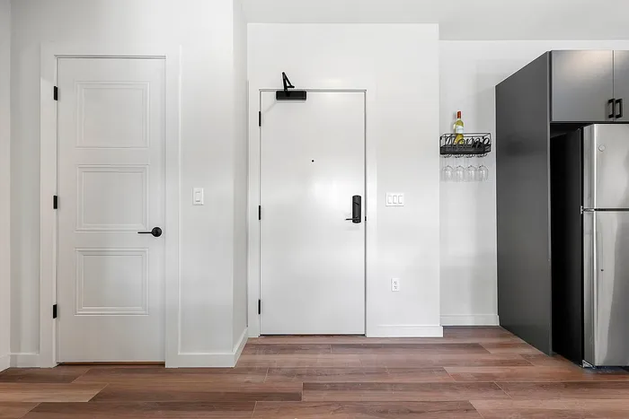 Modern apartment entryway with sleek white doors, elegant wood flooring, and a sophisticated kitchen glimpse with a wine rack.