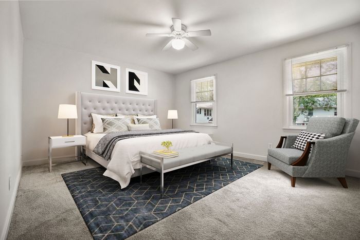 Elegant bedroom with plush gray bed, modern art, cozy armchair, and natural light streaming through large windows.