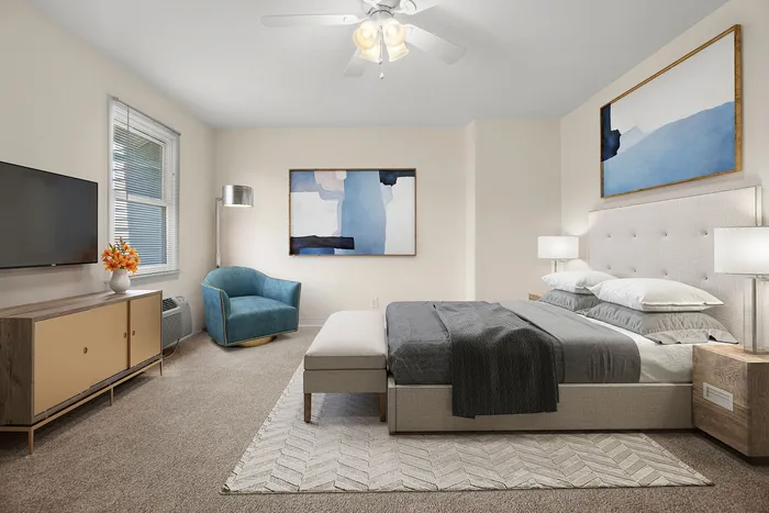 Modern bedroom showcasing a stylish bed with plush pillows, elegant artwork, and a cozy reading chair. Bright natural light accentuates the serene color palette.
