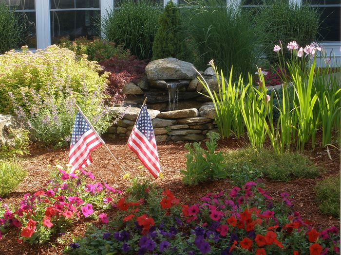 Charming garden oasis featuring vibrant flowers, elegant stone waterfall, and American flags, creating a serene outdoor escape.