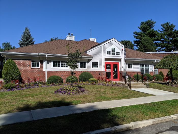 Charming brick building with lush landscaping and vibrant flower beds, featuring a welcoming red door entrance and clear blue skies.