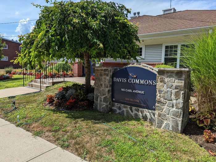 Charming entrance to Davis Commons with lush greenery, vibrant flowers, and a welcoming stone sign at 165 Carl Avenue.