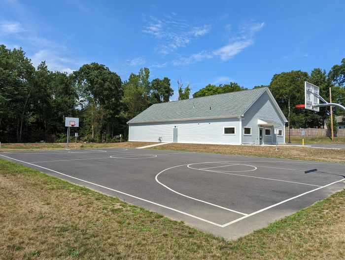 Community sports facility showcasing a pristine basketball court adjacent to a charming clubhouse, surrounded by lush greenery and blue skies.