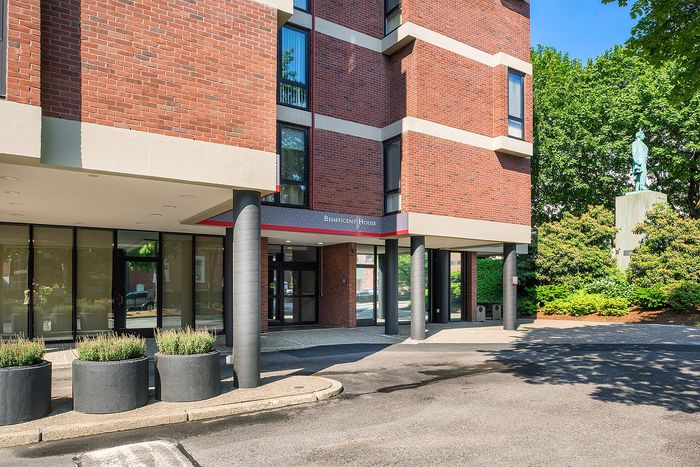 Inviting apartment entrance with modern brick architecture, lush greenery, and ample natural light, ideal for stylish urban living.