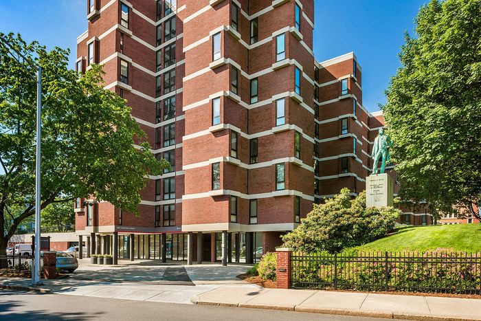Elegant brick apartment building with modern architectural design, surrounded by lush greenery and highlighted by a prominent statue, offering urban living with a touch of serenity.