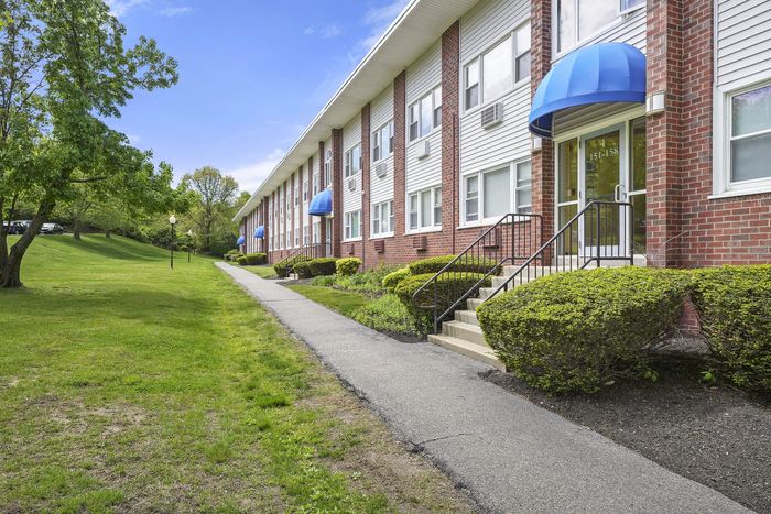 Charming brick apartment building with blue awnings, surrounded by lush greenery and a well-kept lawn, offering a serene living environment.