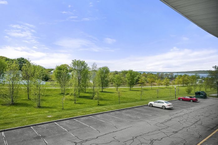Serene outdoor view from apartment balcony showcasing lush green landscape, vibrant trees, and spacious parking area under a clear blue sky.