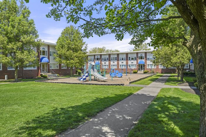 Charming apartment complex with a vibrant playground surrounded by lush greenery and immaculate landscaping under clear blue skies.