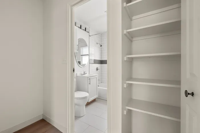 Modern bathroom with sleek white tiles, a stylish round mirror, and an adjacent open shelving unit for ample storage.