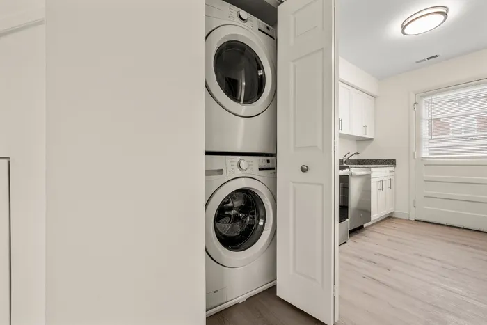 Modern apartment interior with a sleek stackable washer and dryer set in a closet next to a bright kitchen featuring white cabinets and wooden floors.